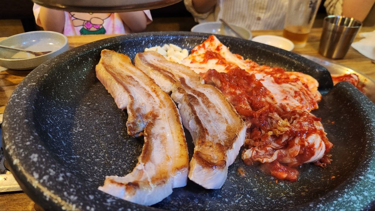 Samgyeopsal browning beside kimchi and garlic on a sloped barbecue pan